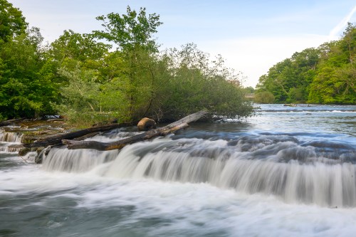 A serene river scene with flowing water over rocks, surrounded by lush green trees and shrubs.