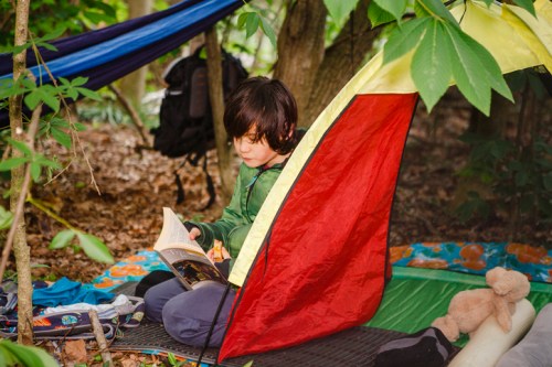 A child reading a book inside a colorful tent in a forested area, surrounded by nature and camping gear.
