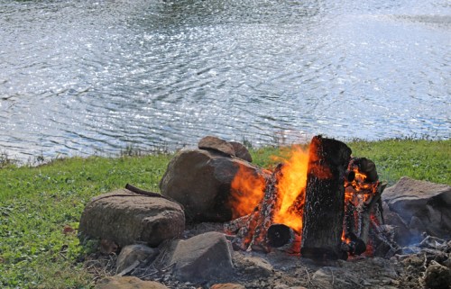 A campfire burns beside a calm lake, surrounded by rocks and green grass under a clear sky.