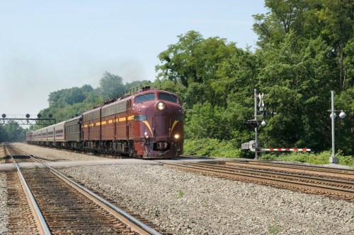 A vintage train approaches a railway crossing surrounded by lush greenery on a sunny day.