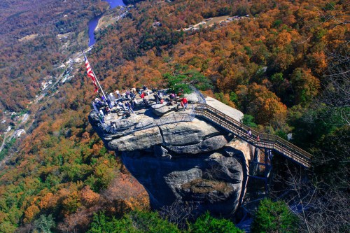 Aerial view of a rocky outcrop with a wooden walkway, surrounded by autumn foliage and a river in the background.