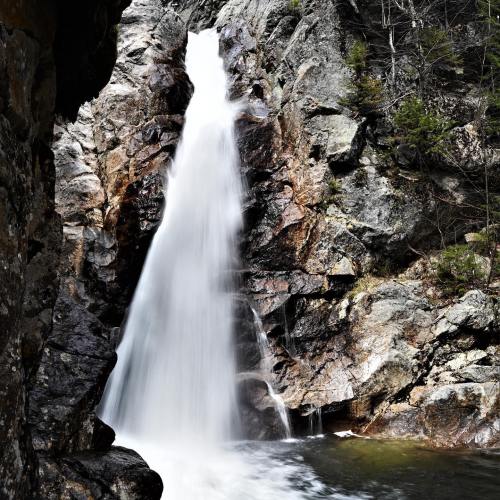 A cascading waterfall flows over rocky cliffs, surrounded by lush greenery and rugged terrain.