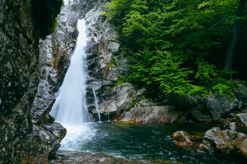 A serene waterfall cascades over rocky cliffs into a tranquil pool, surrounded by lush green trees.