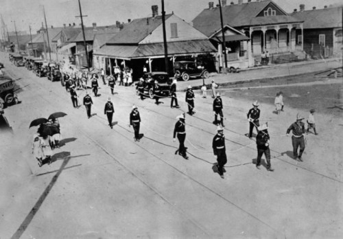 A historic black-and-white photo of a parade with uniformed marchers and spectators along a street.