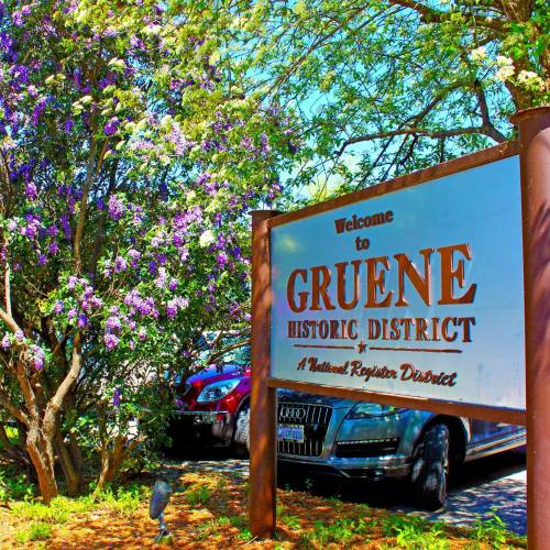 Welcome sign for Gruene Historic District, surrounded by blooming trees and parked cars. Bright, sunny day.