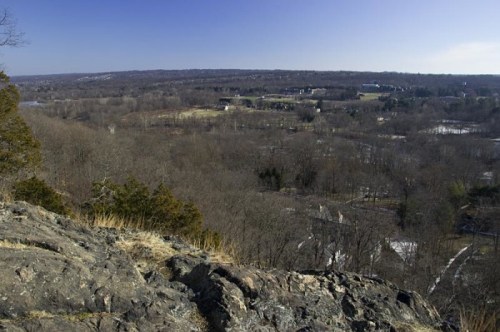 A panoramic view from a rocky outcrop overlooking a vast, wooded landscape under a clear blue sky.