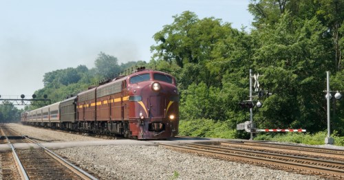 A vintage train approaches a crossing, surrounded by lush greenery and signaling lights in the background.