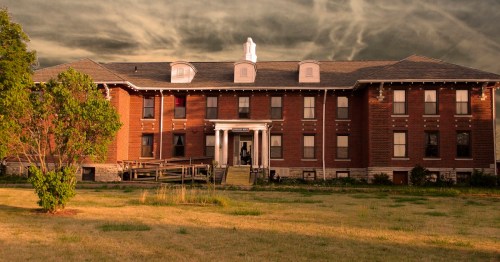A historic brick building with a porch, surrounded by grass, under a dramatic sky.