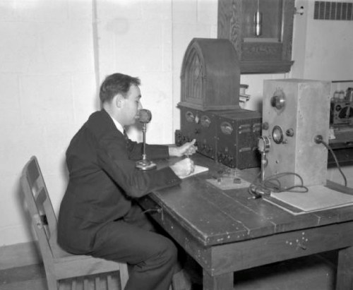 A man in a suit sits at a desk, operating vintage radio equipment and speaking into a microphone.