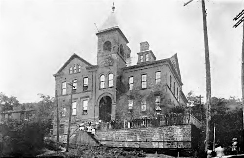 Historic brick building with a tower, surrounded by trees and people, in a black-and-white photograph.