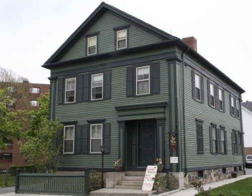 A large green historic house with black shutters and a sign in front, surrounded by greenery and urban buildings.