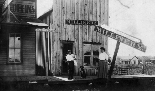 Black and white photo of two women and a dog outside a millinery shop, with wooden buildings and signs in the background.