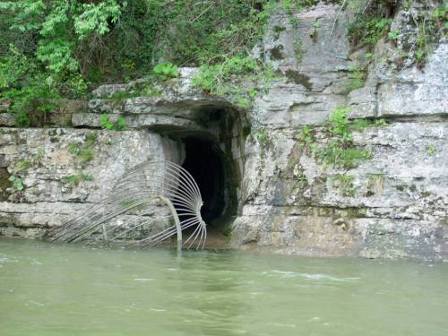 A rocky riverbank with a dark cave entrance and a bent metal structure partially submerged in the water.