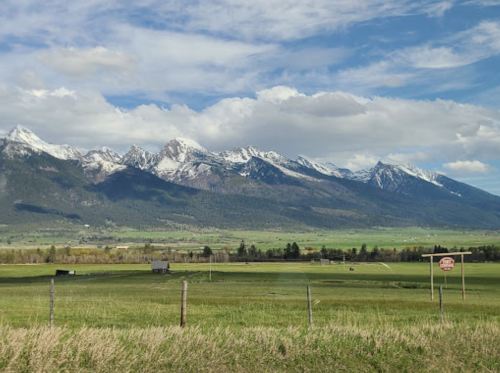 A scenic view of snow-capped mountains under a blue sky, with green fields and a wooden sign in the foreground.