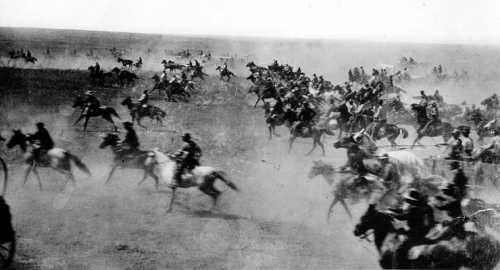 A historic black-and-white photo of a large group of horse riders galloping through a dusty landscape.