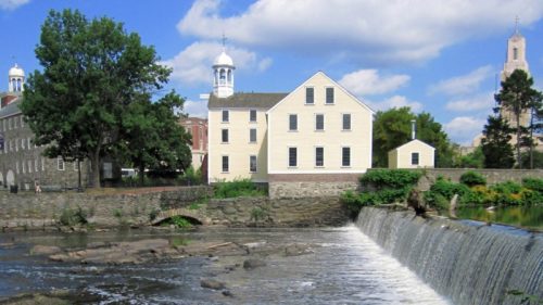 A serene riverside scene featuring a waterfall, historic buildings, and lush greenery under a blue sky with clouds.