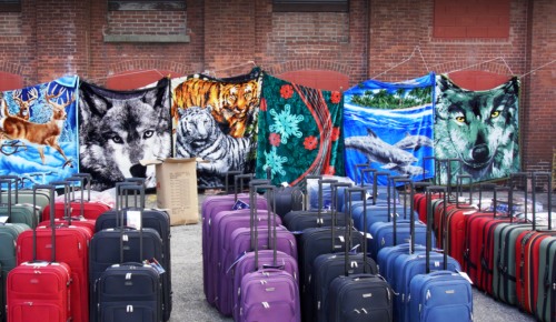 A row of colorful blankets featuring animal designs hangs on a brick wall, with various suitcases in the foreground.