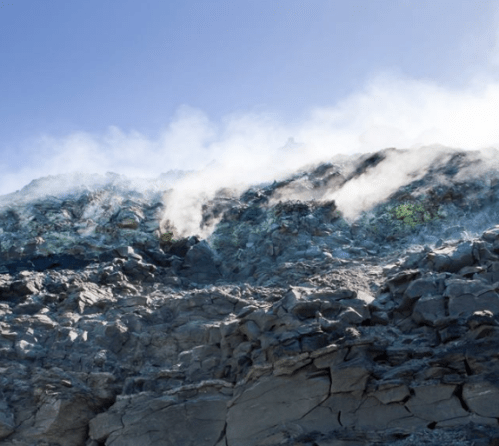 Steam rises from rocky terrain under a clear blue sky, showcasing a rugged, volcanic landscape.