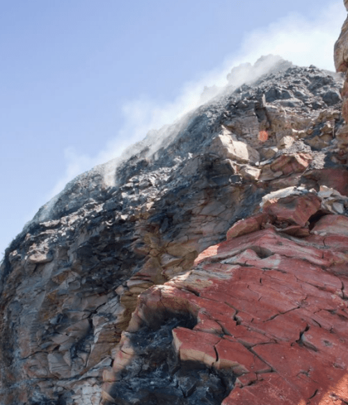 A rocky cliff with layers of red and black stone, emitting wisps of smoke against a clear blue sky.