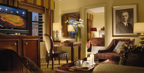 A cozy hotel room with a TV showing a baseball game, a desk, and elegant decor, featuring a portrait on the wall.