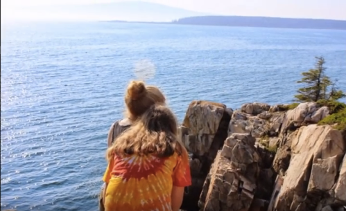 Two people stand on rocky cliffs overlooking a calm ocean, with trees and distant land visible in the background.