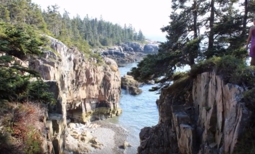 A scenic view of a rocky coastline with trees, featuring a narrow waterway and a person standing on a cliff edge.
