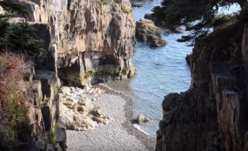 Rocky cliffs frame a serene coastal scene with a pebbled beach and calm blue waters.
