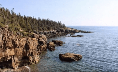 A serene coastal scene with rocky cliffs, calm waters, and a forested shoreline under a clear blue sky.
