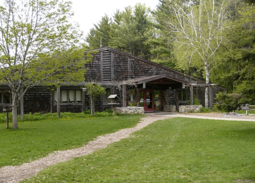 A rustic wooden building surrounded by trees and a grassy area, with a pathway leading to the entrance.