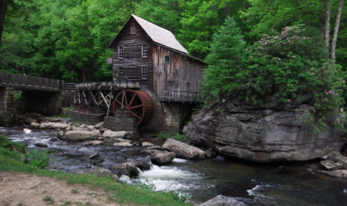 A rustic wooden mill with a waterwheel beside a flowing stream, surrounded by lush green trees and rocky terrain.