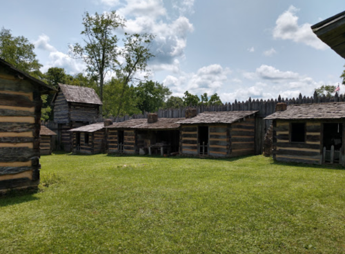 A historic settlement with log cabins and a wooden fence, surrounded by green grass and trees under a blue sky.