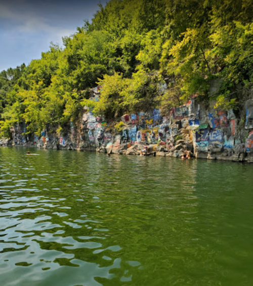 A green lake with a rocky shore covered in colorful graffiti and lush trees in the background.