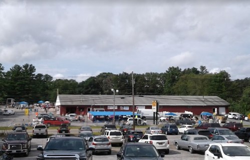 A busy outdoor market scene with numerous cars parked and people gathered near a large red building under a cloudy sky.