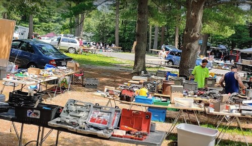 A bustling outdoor flea market with tables full of tools and equipment, surrounded by trees and parked cars.
