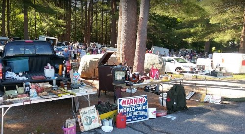 A bustling flea market scene with various items for sale on tables under trees, surrounded by cars and shoppers.