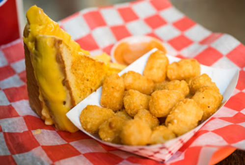 A grilled cheese sandwich with melted cheese and a side of crispy tater tots on a red and white checkered tray.