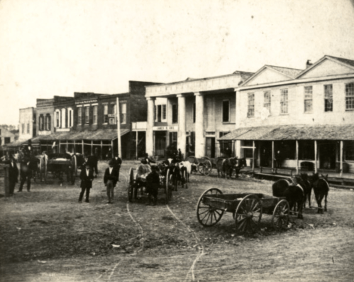 Historic street scene with horse-drawn carriages, buildings, and people in early 20th-century attire.