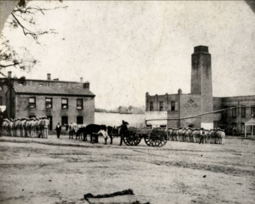 Historic black-and-white photo of a group of people in striped uniforms, a horse-drawn cart, and buildings in the background.