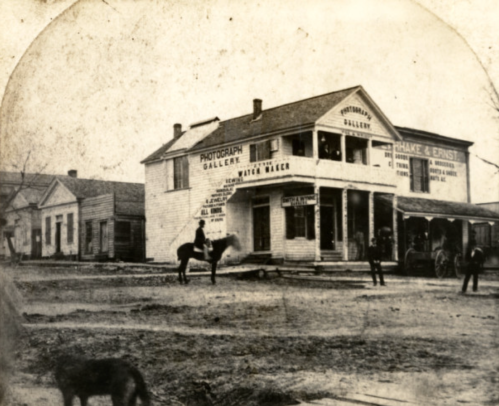 Historic photograph of a photography gallery with people and horses outside, set in a dusty, old town.