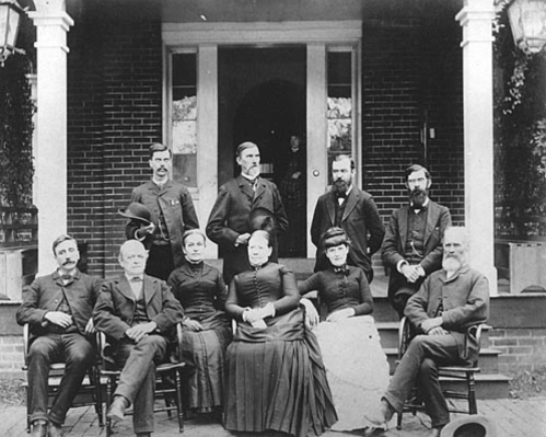 A historic black-and-white photo of a group of men and women posed on a porch, dressed in 19th-century clothing.