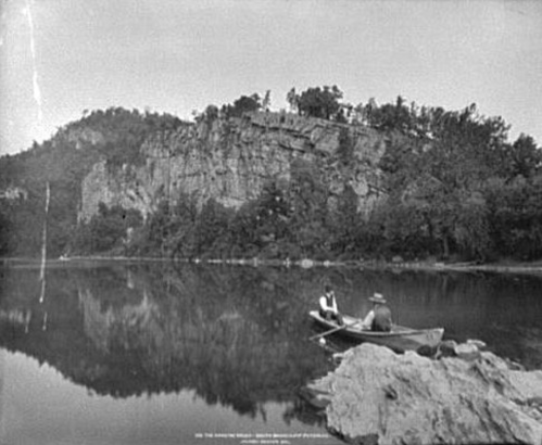 Two people in a small boat fishing on a calm lake, surrounded by rocky cliffs and trees in the background.