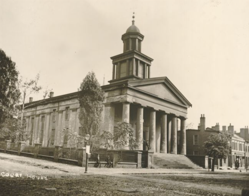 Historic courthouse building with tall columns and a clock tower, surrounded by trees and a street scene.
