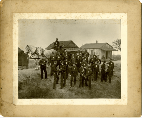 A historical group photo of soldiers in uniform, standing with a horse near wooden buildings.