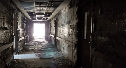 A dimly lit, abandoned hallway with peeling walls and debris scattered on the floor, leading to a bright exit.