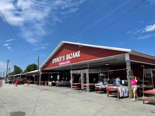 A red-roofed market building with "Spence's Bazaar" sign, featuring outdoor vendor stalls and shoppers on a sunny day.