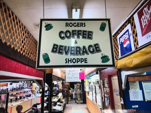 Sign for "Rogers Coffee & Beverage Shoppe" hanging in a market, with various beverage logos visible in the background.