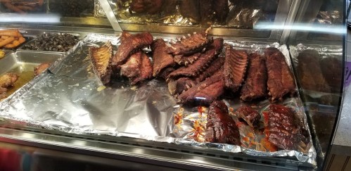 A display of grilled ribs on a silver tray, glistening with barbecue sauce, in a food service setting.