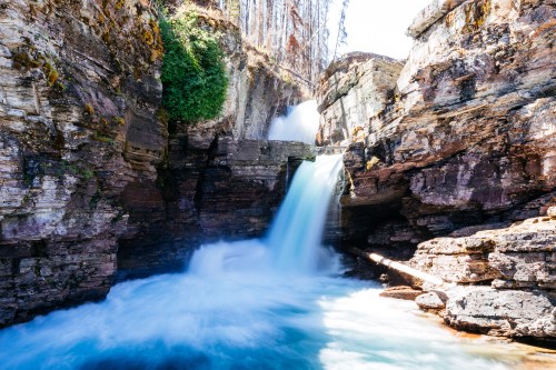A serene waterfall cascades over rocky cliffs into a clear blue pool, surrounded by lush greenery and sunlight.