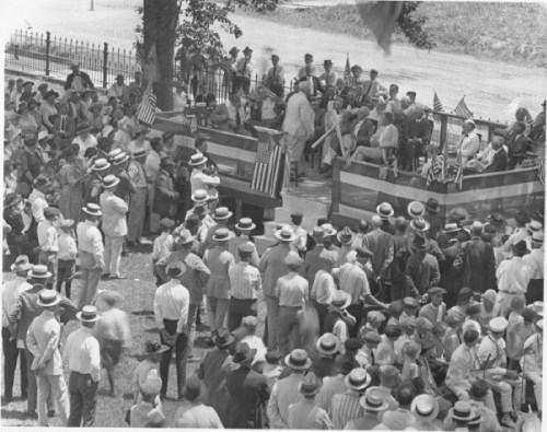 A large crowd gathers outdoors, listening to speakers at a podium, with flags and trees in the background.