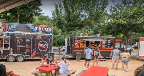 Two food trucks in a park setting, one serving tacos and the other offering various dishes, with people dining at picnic tables.
